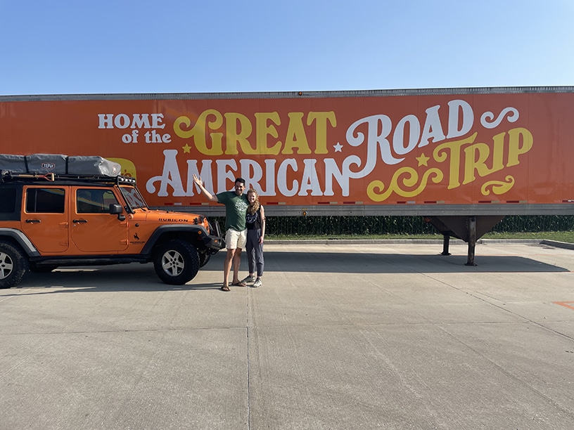 Two people standing in front of a semi truck that says "Home of the Great American Road Trip" with an orange jeep off to the side